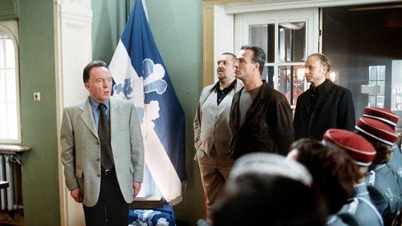 Drei Männer betreten einen Raum mit einer blau-weißen Standarte und einer Reihe von uniformierten Personen und schauen sich um.