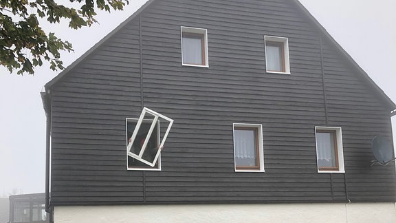 Die Front eines zweistöckigen Hauses, in der ersten Etage bringt jemand einen Fensterladen vor einem Fenster an.
