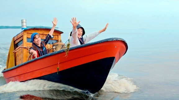 Pertsa (Olavi Kiiski) and Kilu (Oskari Mustikkaniemi) winken Kapitän Ritavuori (Veeti Kallio) aufgeregt von ihrem Boot aus zu.