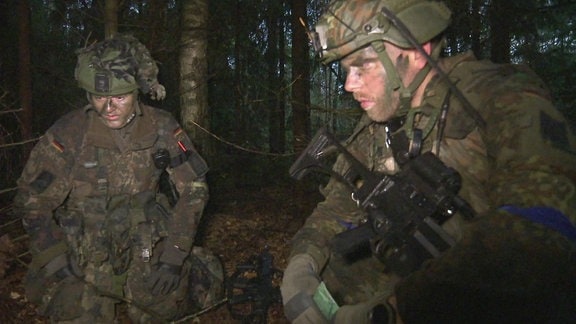 Zwei junge Männer in Uniform und mit Waffen hocken in einem dunklen Wald.