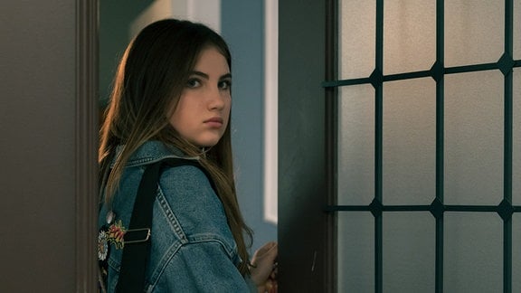 Eine junge Frau schaut durch eine offene Tür zum Betrachter.