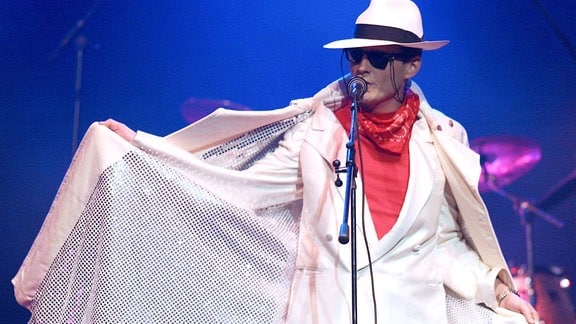 Falco (Manuel Rubey) steht in einem weißen Umhang auf der Bühne am Mikrophon.