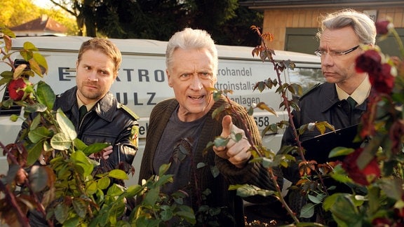 Frido Schulz (Robert Atzorn) mit seinem Sohn David (Janek Rieke, li.) und einem Kollegen hinter einem Rosenbusch.