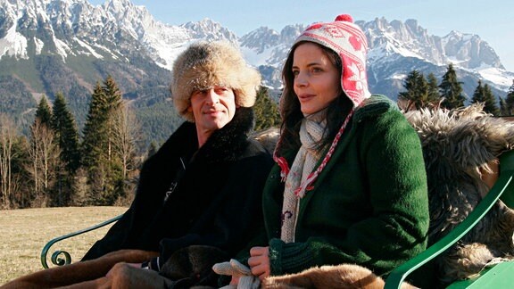 Lisa (Elisabeth Lanz, re.) spricht und der Baron von Lien (Max Tidof) sieht sie aufmerksam an. Sie sitzen draußen, im Hintergrund sind die Berge zu sehen. 