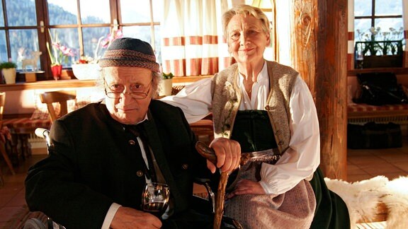 Der Dorfarzt Felix (Kurt Weinzierl) blickt sich an einem Gehstock festhaltend in die Kamera, in der anderen Hand hat er ein Weinglas. Irmi (Ingrid Burkhard) sitzt hinter ihm und lächelt in die Kamera.
