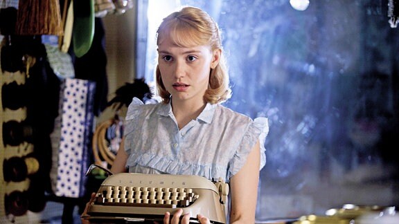 Ein junge Frau trägt eine Schreibmaschine und schaut ängstlich.