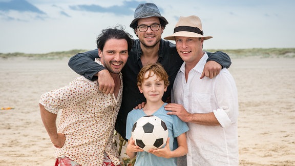 Drei Männer stehen um einen Jungen am Strand der einen Fußball hält. Die Stimmung ist locker gelöst.