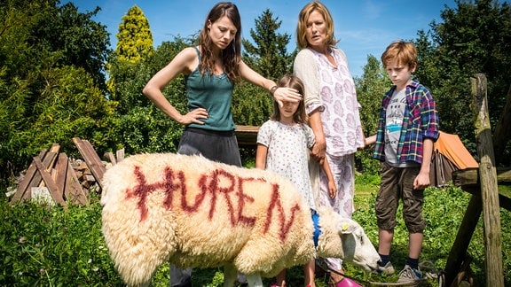 Eine Frau mittleren Alters, eine junge Frau und zwei Kinder stehen hinter einem angebundenen Schaf, auf dessen Fell mit roter Schrift "Huren" steht. Die junge Frau hält einem der Kinder die Augen zu.