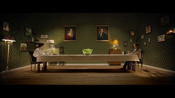 Zwischen ihr (Dorothea Arnold) und ihm (Christian Heiner Wolf) steht der scheinbar unerreichbare Salat auf der Mitte eines langen Tisches. Beide sitzen weit voneinander entfernt an diesem Tisch in einem kahl wirkenden Raum.