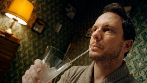 Er (Christian Heiner Wolf) an einem Ende des Esstisches, mit Hilfe eines Strohhalmes aus einem Glas trinkend.