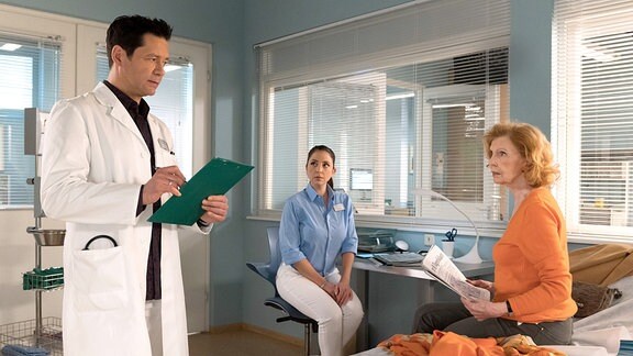Ein Arzt im weißen Kittel steht, zwei Frauen sitzen in einem Raum.
