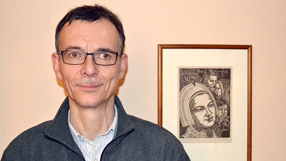 Ein Mann mit kurzen Haaren und Brille steht vor einer Wand. Neben ihm hängt ein Bild mit einer abgebildeten Frau in sorbischer Tracht.