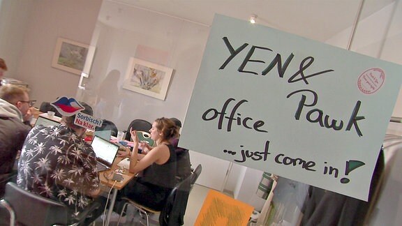 Menschen sitzen hinter einer Glastür an einem Konferenztisch. An der Tür ein Zettel: "YEN and Pawk office ... just come in !"