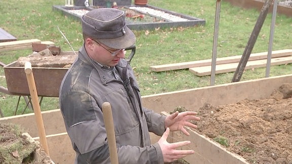 Ein Mann steht auf einem Friedhof in einem leeren Grab. Im Hintergrund eine Schubkarre.