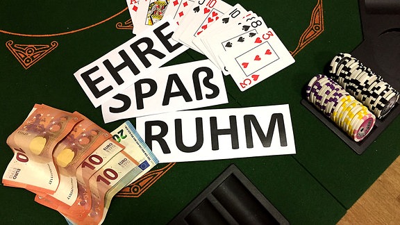 Auf einem Pokertisch liegen Geldscheine, daneben Schilder mit Aufschrift "Ehre", "Spaß", "Ruhm" und Spielkarten sowie Pokerchips