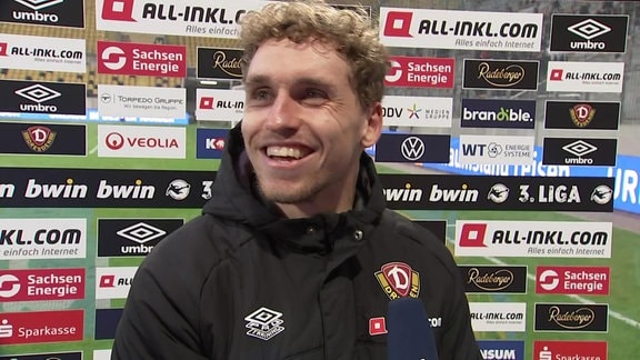 Dynamo-Spieler Luca Herrmann grinsend vor einer Werbetafel im MDR-Interview