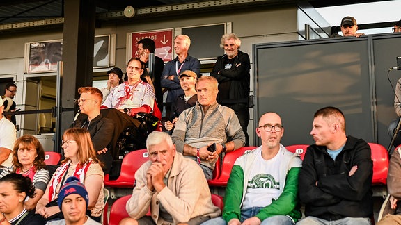 Bundestrainer Christian Wück (Mitte, in grauer Strickjacke) weilt unter Zuschauer*innen auf einer Tribüne.