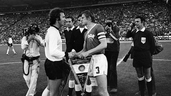 Die Spielführer Franz Beckenbauer (BRD, li.) und Bernd Bransch (DDR) tauschen die Wimpel vor Schiedsrichter Ramon Barreto (Uruguay, Mitte)