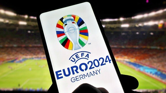  Das Logo der UEFA Euro 2024 ist auf einem Smartphone vor einem Stadionhintergrund zu sehen.