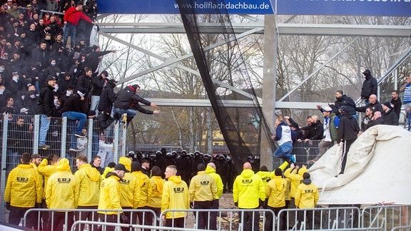 Eine Gruppe Polizist*innen im Hintergrund von Ausschreitungen auf den Zuschauerrängen eines Fußballspiels
