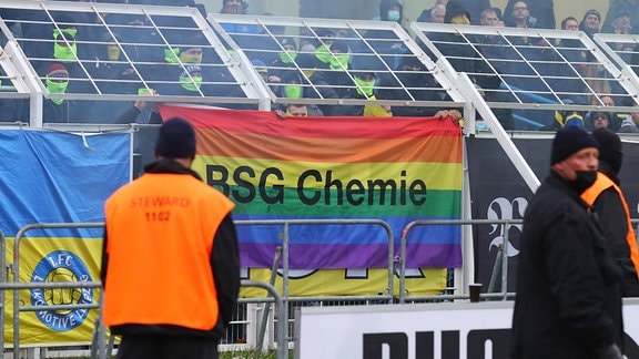 Fans von Lokomotive Leipzig befestigen eine BSG Chemie - Regenbogen Fahne am Stadionzaun.