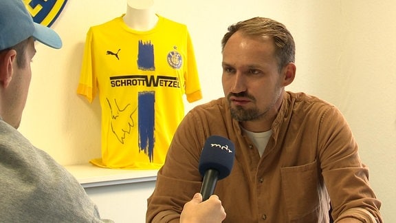 Sportlicher Leiter in einem Büro im Interview, im Hintergrund ein blau-gelbes Trikot des 1. FC Lok Leipzig.