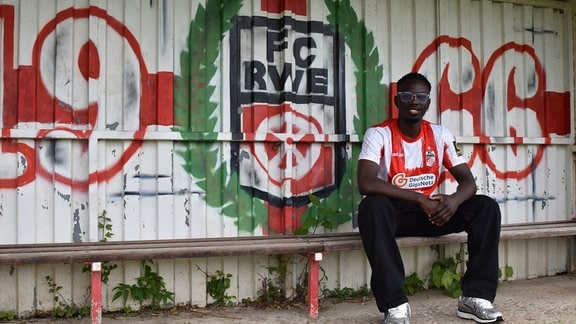 Neuzugang Yesua Motango sitzt auf einer Bank vor einer Wand, auf die das Logo von FC Rot-Weiß Erfurt gesprüht ist.