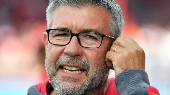 Urs Fischer - Trainer von Union Berlin. Ein Mann greift sich mit der linken Hand an das linke Ohr. Er trägt Bart und Brille.