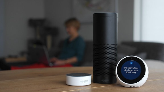 Ein schwarzes Echo 1 Gerät neben einem weißen Echo Spot und einem weißen Echo Dot 2 und von Amazon steht auf einem Tisch in einem Wohnzimmer. Auf dem Bildschirm des Echo Spot ist das Logo von MDR THÜRINGEN Das Radio zu sehen.