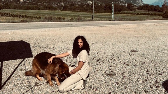 Eine junge Frau mit langem schwarzen Haar Kniet am Straßenrand vor einem Hund. Im Hintergrund sind Berge zu sehen.