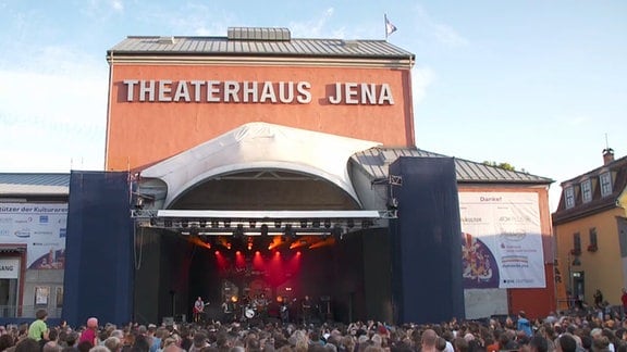 Eine Menschenmenge steht vor einem roten Gebäude mit der Aufschrift "Theaterhaus Jena"