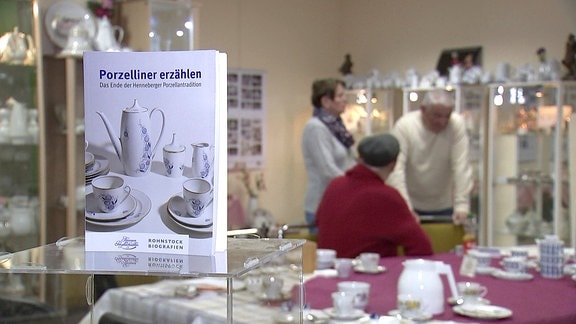 Buch "Porzelliner erzählen" vor einem Tisch mit Porzellan und drei Menschen  im Gespräch