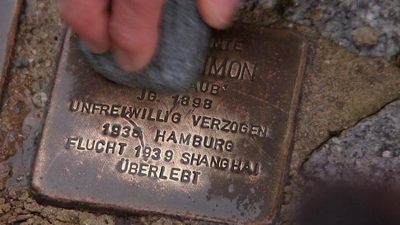 Drei Finger halten einen Schwamm, mit dem ein Stolperstein geputzt wurde. Auf dem Storperstein ist zu lesen: "unfreiwillig verzogen/ 1938 Hamburg /Flucht 1939 Shanghai / überlebt"