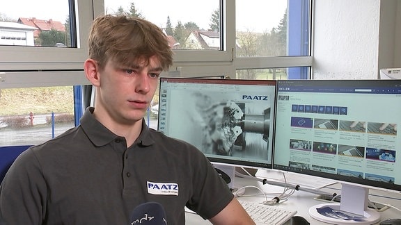 Ein Azubi der Paatz-Viernau GmbH sitzt mit Unternehmens-T-Shirt am Schreibtisch, im Hintergrund zwei Monitore mit Bildern des Unternehmens