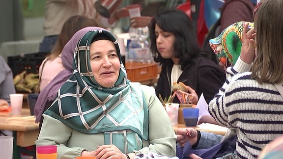 Eine muslimische Frau mit Kopftuch sitzt an einem Tisch. im Hintergrund Frauen, die Essen