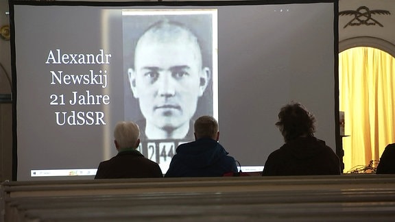 drei Menschen von hinten, die in einer Kirchenbank sitzen und auf ein projeziertes Bild eines junge Soldaten aus der UdSSR schauen 