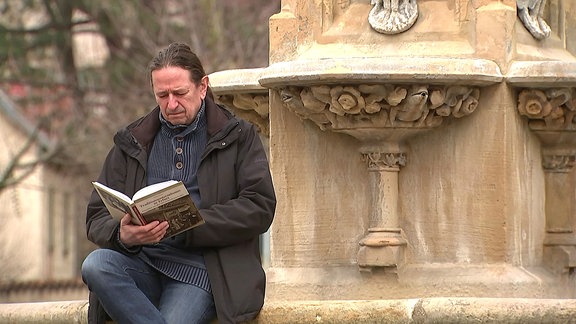 Der Autor Ulrich Seidel sitzt an einem Brunnenrand in Erfurt und hält sein Buch "Traditionsunternehmen in Erfurt" in der Hand