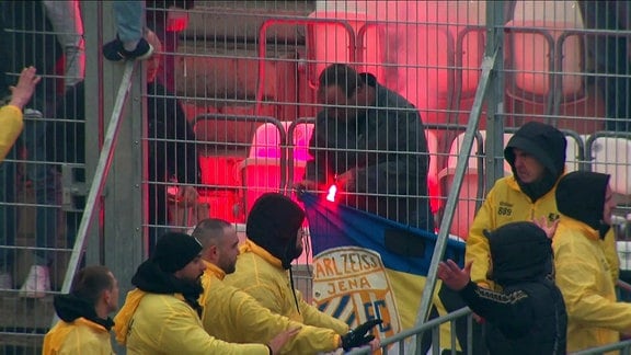 Mehrere Ordner in gelben Jacken stehen vor einem Schutzgitter auf einer Stadion-Tribüne, im Hintergrund zündet ein Mann ein rotes Bengalow an