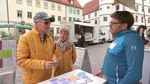Ein Mann und eine Frau stehen auf dem Marktplatz in Hildburghausen vor dem Info-Tisch der Verbraucherzentrale und sprechen mit einem Mitarbeiter der Verbarucherzentrale 