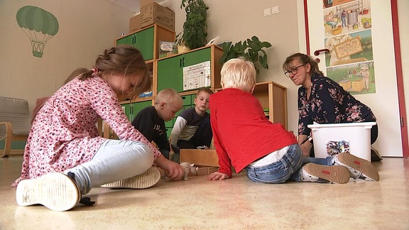 Vier Kindergartenkinder und eine Erzieherin knien auf dem Erdboden und spielen mit Spielzeugtieren 