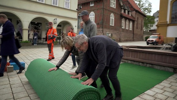 Drei Menschen rollen in der Innenstadt von Suhl einen großen grünen Teppich aus