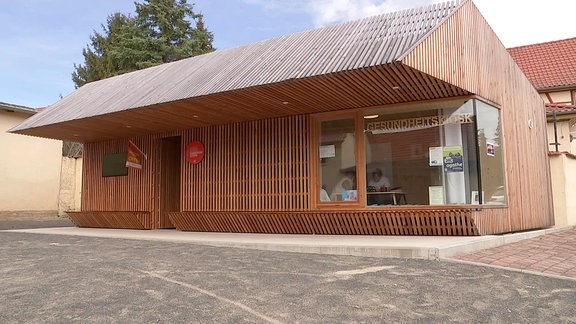 Ein Gebäude aus Holz mit großem Glasfenster, auf dem "Gesundheitskiosk" steht 