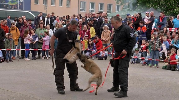 Zwei Polizisten führen mit einem Hund einer Menschenmenge etwas vor.