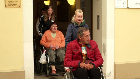 Zwei junge Mädchen schieben zwei Menschen im Rollstuhl durch eine Tür