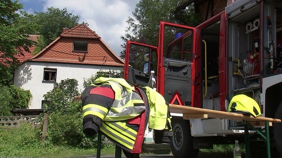 Ein Feuerwehrauto und eine Feuerwehrjacke vor einem Haus mit ausgebrannten Fenstern 
