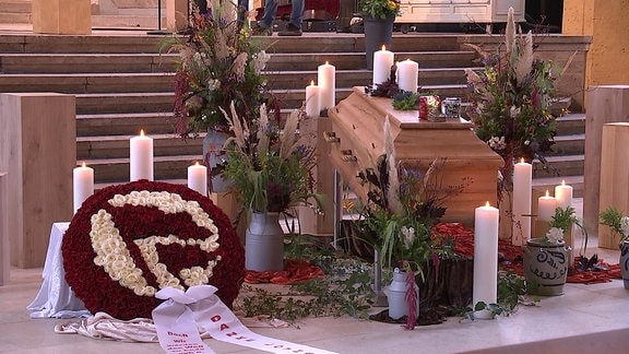 in einer Kirche steht ein mit Kerzen und Blumen geschmückter Sarg, davor ein aus Blumen gestecktes Antifa-Zeichen