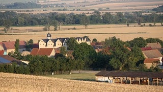 Blick auf ein Dorf, das von Getreidefeldern umgeben ist. In der Mitte des Ortes steht ein großes Schloss.
