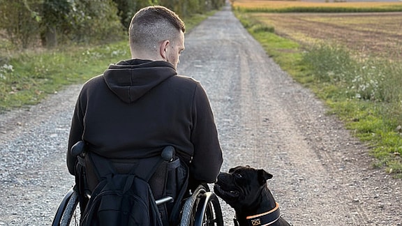 Ein Hund sitzt neben einem Rollstuhlfahrer auf einem Feldweg