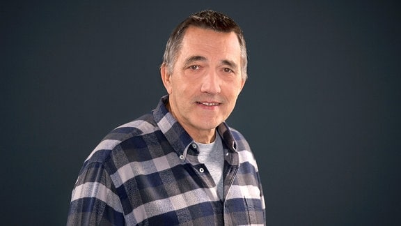 Peter Zudeick, ein Mann mit dunklen Haaren und einem karierten Hemd vor einem schwarzen Hintergrund