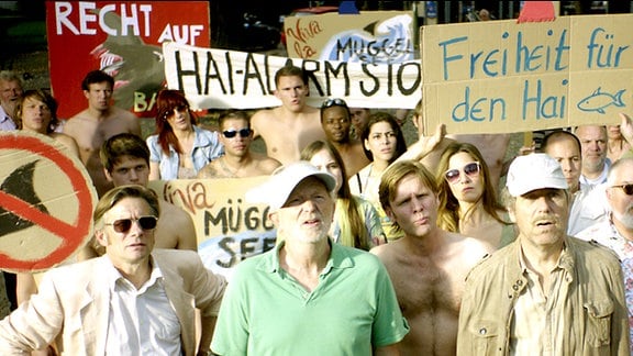 Eine Szene aus dem Film "Hai-Alarm am Müggelsee": Menschen mit Pappschildern bei einer Hai-Demo.